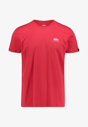 Czerwona koszulka z krótkim rękawem i okrągłym dekoltem, z małym białym logo Alpha Industries na lewej piersi oraz czarną metką na lewym rękawie.
