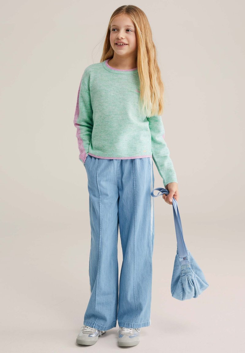 Pull vert menthe avec des accents roses, associé à un pantalon large bleu clair et un petit sac bleu. La tenue présente des textures douces et un design décontracté.