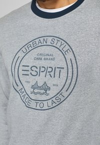 Sweat-shirt gris avec un design rond bleu marine. Le texte comprend "ESPRIT" et "URBAN STYLE", avec un graphisme de pont et de vagues. Col bleu marine.