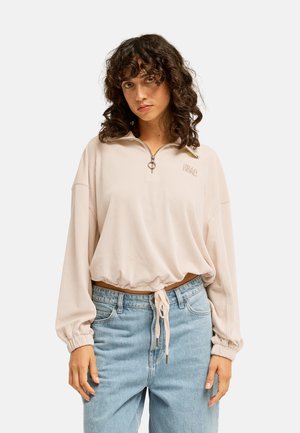 Frau mit lockigem Haar, die einen hellbeigen Reißverschluss-Pullover mit Kordelzug in der Taille und hellblaue hochtaillierte Jeans vor einfarbigem Hintergrund trägt.