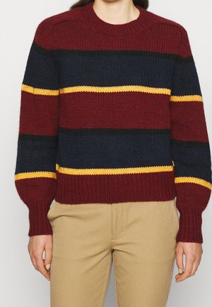 Personne portant un pull en tricot avec des rayures horizontales bordeaux, marine, noir et jaune, associé à un pantalon beige.