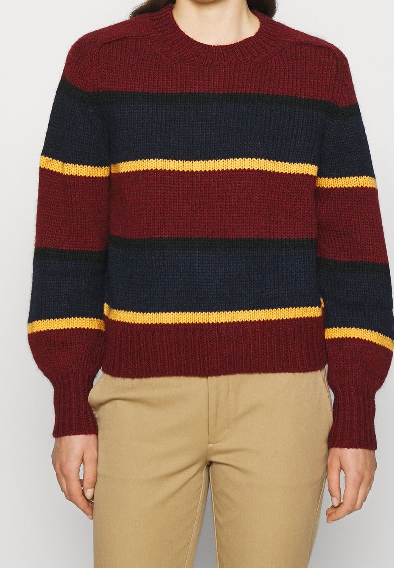 Personne portant un pull en tricot avec des rayures horizontales bordeaux, marine, noir et jaune, associé à un pantalon beige.