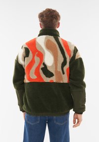 Chaqueta de forro polar en bloques de color verde y naranja que cuenta con un cuello alto, tejido texturizado y un diseño de corte relajado con puños elásticos.