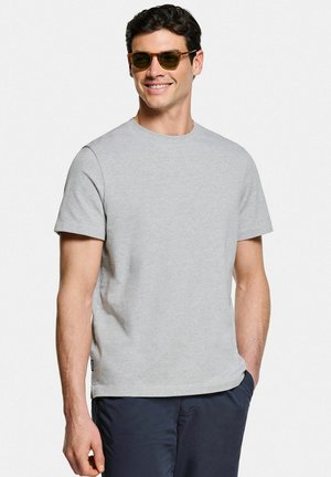 Homme portant des lunettes de soleil marron, un t-shirt gris clair à manches courtes et un pantalon foncé, souriant avec une main dans la poche sur fond blanc.