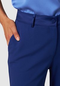 Personne portant un pantalon bleu cintré avec un bouton assorti et une passoire de ceinture, la main reposant dans la poche latérale.