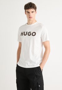 Fehér pamut póló, a mellkason feltűnő fekete "HUGO" felirattal. A modellen fekete cargo nadrág van, kezét a zsebébe dugta.