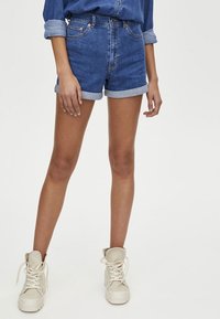Shorts en denim de bleu moyen avec des ourlets roulés, associés à des baskets montantes beiges. Le tissu semble robuste, avec un design classique à cinq poches.