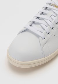 Sapatilha de couro azul claro com atacadores brancos, bico arredondado e sola de borracha creme. Apresenta detalhes cosidos e pequenas perfurações nas laterais.