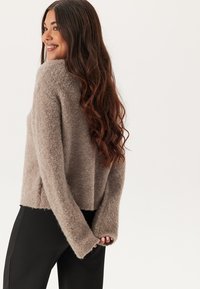 Maglione beige peloso con una vestibilità comoda, spalle scese e maniche lunghe; abbinato a pantaloni neri, esposto su uno sfondo neutro.