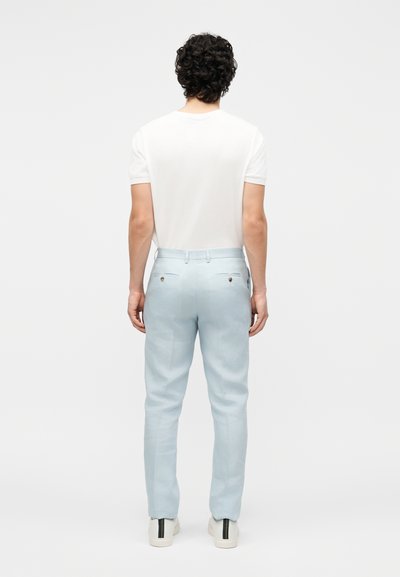 Άνδρας με σγουρά μαλλιά, που φοράει λευκό κοντομάνικο πουκάμισο, ανοιχτό μπλε παντελόνι και λευκά αθλητικά, στέκεται με την πλάτη σε μονόχρωμο φόντο.