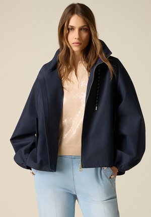 Jonge vrouw met los golvend haar draagt een oversized donkerblauwe jas over een beige top met pailletten en lichtblauwe spijkerbroek, handen in de zakken.