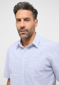 Jasnoniebieska koszula z krótkim rękawem o teksturowanym wzorze, z kołnierzykiem zapinanym na guziki oraz pojedynczą kieszenią na piersi. Posiada zaokrąglony dół oraz beżowe guziki.