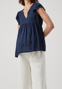 Navy blue blouse with short ruffled sleeves and a V-neckline, featuring a gathered waistline. Paired with loose, light-colored trousers.