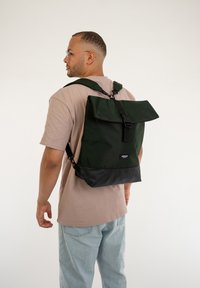 Zelený plátěný batoh s překlápěcím víkem, nastavitelnými popruhy a černou spodní částí. Na přední straně se nachází logo.