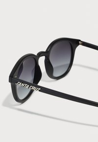 Schwarze Sonnenbrille mit Verlaufsgläsern. Verfügt über ein rundes Design und das "SANTA CRUZ"-Logo in Silber am Bügel für zusätzliche Details.