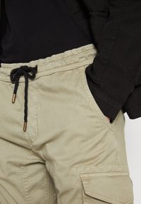 Knälånga, khaki cargo-shorts med elastisk midja och svart dragsko. Har sidfickor och en strukturerad tygfinish.