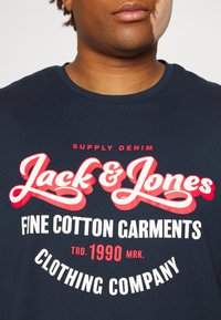 Mornarsko modra bombažna majica s poudarjenim rdečim in belim grafičnim besedilom, ki vključuje ime blagovne znamke "Jack & Jones" in "Fine Cotton Garments."