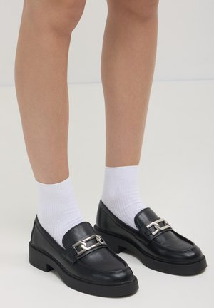 Mocassins en cuir noir avec une semelle épaisse, ornés d'un accent de chaîne en argent. Portés avec des chaussettes blanches à mi-mollet, montrant une texture lisse et un design robuste.