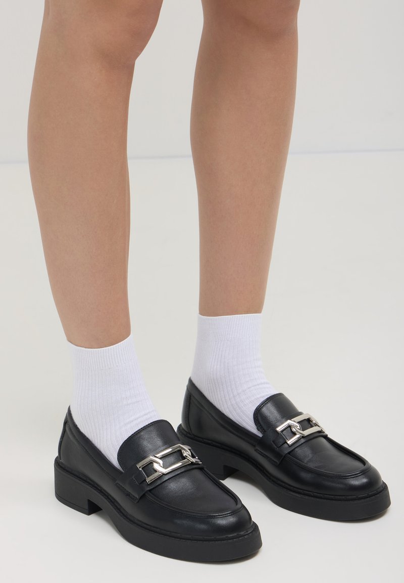 Mocassini in pelle nera con suola spessa, caratterizzati da un accento in catena argentata. Indossati con calzini bianchi alla caviglia, mostrando una texture liscia e un design robusto.