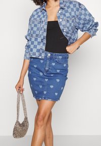 Light blue denim jacket with a checkered pattern, paired with a light blue denim skirt featuring heart patterns. Holding a silver beaded bag.