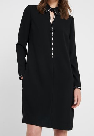 Frau trägt ein schwarzes langärmeliges Kleid mit einem mit Nieten besetzten Kragen, einer vertikalen Nietenreihe vorne und Nieten an den Manschetten, steht mit teilweise in den Taschen steckenden Händen.