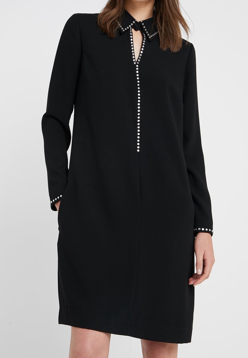 Femme portant une robe noire à manches longues avec un col clouté, une rangée verticale de clous sur le devant et des clous sur les poignets, debout les mains partiellement dans les poches.