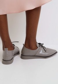 Chaussures en cuir verni gris avec un bout pointu, des lacets noirs et des perforations décoratives, sur un fond clair.