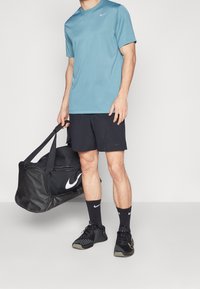 Helles blaues Sportshirt, schwarze Shorts, schwarze Sneakers mit Akzenten, schwarze Duffel-Tasche mit weißem Logo, getragen vor einem neutralen Hintergrund.