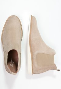 Pár béžových semišových Chelsea bot s elastickými bočními panely a bílými podešvemi, zobrazených z vrchu a ze strany na bílém pozadí.