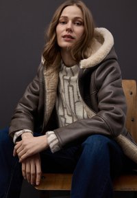 Brun läderjacka med krämfärgad, plyschig shearlingkrage, kombinerad med en mönstrad tröja i kräm och brunt, bärs tillsammans med blå jeans.