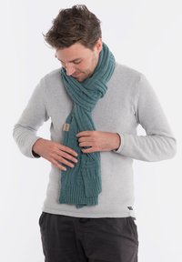 Turkusowy, dziergany szalik o teksturowanym wzorze, z wyraźnym ściągaczem na końcach i małą metką. Noszony luźno na jasnoszarym swetrze.