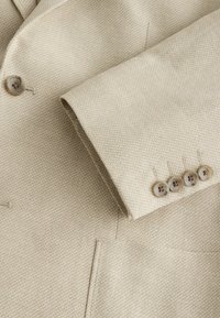 Bézs texturált öltönykabát ujj, négy barna gombbal, amelyek átfedik a fő részt, egy gombbal és egy zsebbel látható.