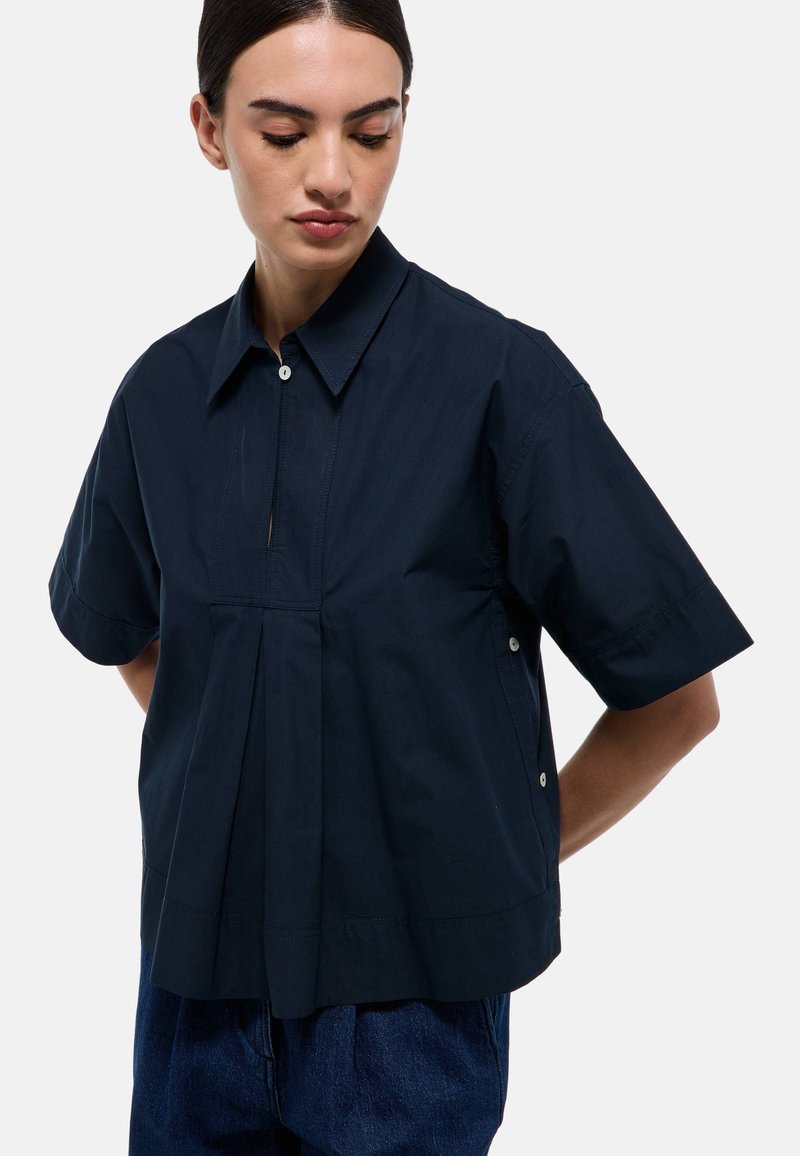 Chemise bleu marine à manches courtes avec boutons, présentant un devant plissé, un col classique et des boutons-pression sur les coutures latérales. Texture de tissu lisse.