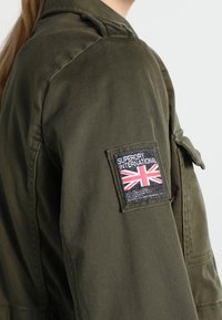 Oliwkowa kurtka z naszywką przedstawiającą brytyjską flagę Union Jack oraz tekst. Tkanina ma gładką teksturę, z klasycznym militarnym wzornictwem i kieszeniami na ramionach.