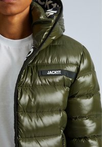 Lähikuva henkilöstä, joka on pukeutunut oliivinvihreään kiiltävään untuvatakkiin, jossa on "JACK1T" -merkki, ja valkoisen paidan alla.