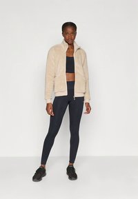 Beige sherpa jas met rits en twee voorzakken, gecombineerd met zwarte legging en sportschoenen, met een aansluitend ontwerp.