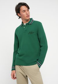 Vihreä pitkähihaisella polo-paidalla, jossa on ribbauskaulus mustavalkoisilla stripeillä. Vasen rinta-logokohta mustana. Sileä puuvillamateriaali.