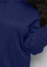 Pull bleu marine avec un corps lisse et des manches à texture côtelée, conçu avec un col montant et des poignets ajustés.