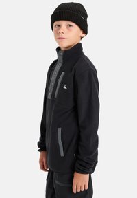 Schwarze Fleecejacke mit grauen Akzenten, hohem Kragen, Druckknöpfen und seitlichen Taschen. Kombiniert mit einer schwarzen Strickmütze. Strukturierter Stoff.