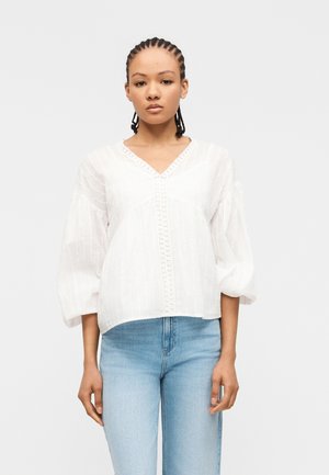 Femme aux cheveux tressés portant un chemisier blanc texturé avec bordure en dentelle et un jean bleu clair, debout devant un fond blanc uni.