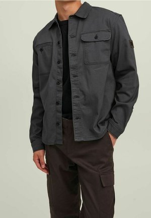 Homme portant une surchemise gris foncé à boutons avec des poches à rabat sur une chemise noire et un pantalon cargo foncé.