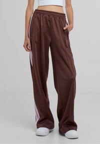 Pantalones atléticos de pierna ancha color marrón con cintura elástica, bolsillos laterales y rayas rosas y blancas a lo largo de los costados. Textura de tela suave.