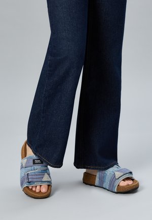 Füße, die blaue, gemusterte Sandalen mit offenen Zehen, verstellbaren Riemen und Korksohlen tragen, kombiniert mit dunkelblauen weiten Jeans.