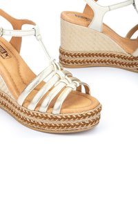 Sandales en wedge métalliques dorées avec plusieurs sangles, semelle intérieure tissée et semelle en jute tressé. Doublure en cuir souple pour le confort.