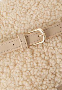 Μπεζ υφασμάτινη επιφάνεια με υφή, απαλή και βελούδινη, συμπληρωμένη από μια ανοιχτή μπεζ δερμάτινη λωρίδα με χρυσή μεταλλική πόρπη.