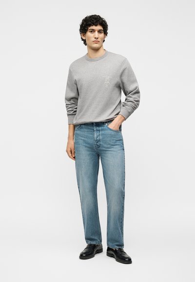 Άνδρας με σγουρά μαλλιά που φοράει γκρι φούτερ, ανοιχτό μπλε τζιν και μαύρα δερμάτινα παπούτσια, στέκεται με το ένα χέρι στην τσέπη.