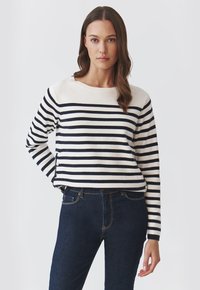 Sweter w paski w kolorze kremowym i granatowym, z okrągłym dekoltem, długimi rękawami i detalem w postaci guzika z boku, zestawiony z ciemnoniebieskimi dżinsami.
