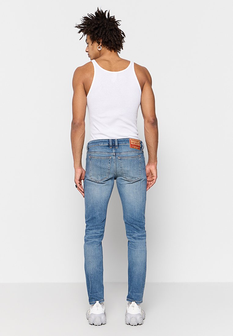 Jean en denim bleu clair avec une coupe slim, présentant une poche arrière en patch et une étiquette de marque au-dessus de la ceinture. Porté avec un débardeur blanc.