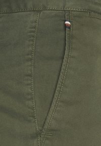 Olijfgroene stof met een gladde textuur, voorzien van contrasterende stiksels en een kleine gestreepte accent bij de zak.