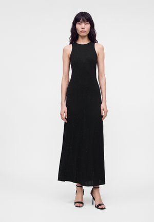 Donna con lunghi capelli neri ondulati che indossa un vestito nero senza maniche lungo fino alla caviglia e sandali neri con tacco alto, in piedi davanti a uno sfondo semplice.